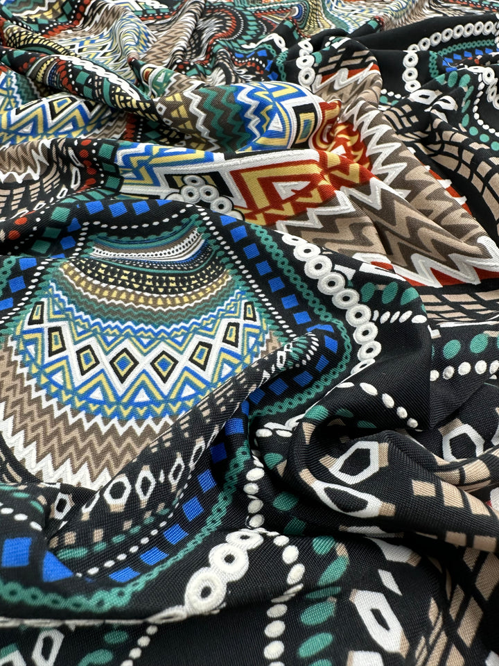 A close-up photo of a multi-use fabric featuring an intricate, colorful, geometric pattern. The design includes various shapes such as diamonds and zigzags in shades of blue, green, red, brown, and white, flowing across a black background. The Textured Lycra - Africa - 150cm by Super Cheap Fabrics has a smooth texture and is draped loosely.