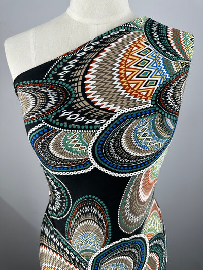 A close-up view of a mannequin wearing a one-shoulder garment made from Super Cheap Fabrics' Textured Lycra - Africa - 150cm, adorned with an intricate, multicolored tribal pattern. The design features overlapping, elongated oval shapes with geometric motifs, including diamonds and zigzags, in shades of black, white, green, brown, and blue.