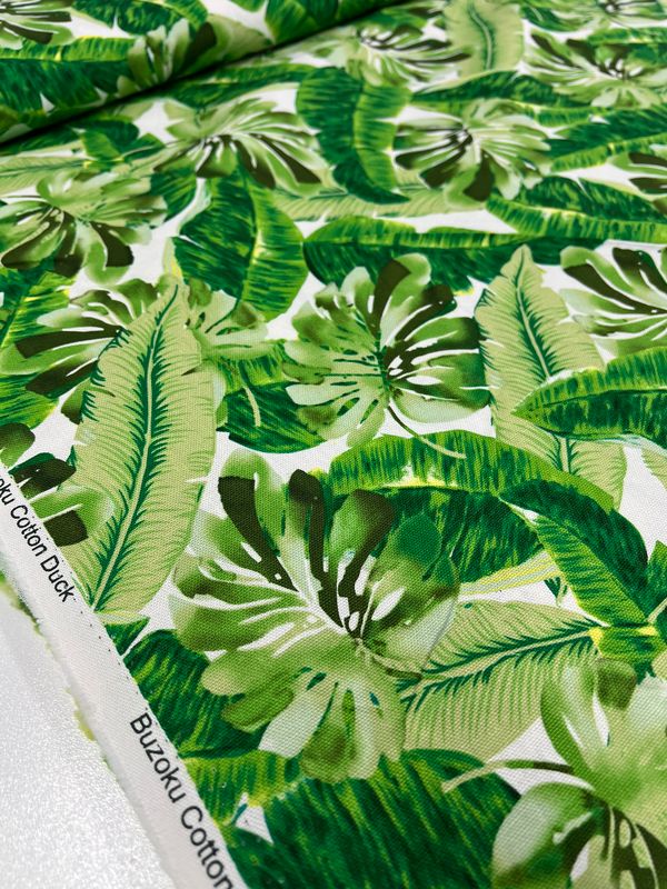 A close-up of the Printed Canvas - Monsterra (112cm) by Super Cheap Fabrics showcases its tropical pattern with lush green banana and palm leaves, ideal for tote bags or home décor. The edge shows text: "Buzoku Cotton Duck," with vibrant colors enhancing the design.