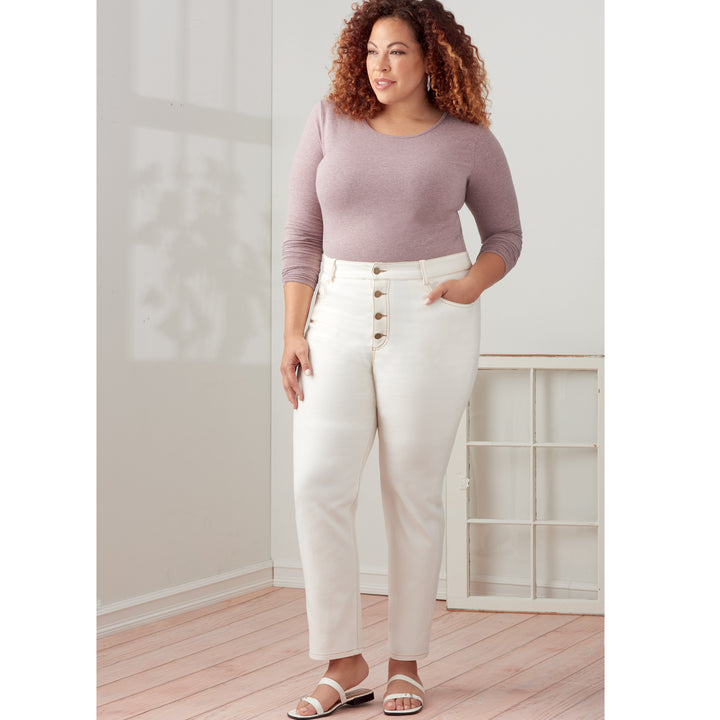 A woman stands indoors wearing a lavender long-sleeve top and high-waisted, ankle-length white pants with buttons, made using the "Simplicity - S9266 - Misses and Women's Vintage Jeans" pattern from Super Cheap Fabrics. She has one hand in her pocket, wears white sandals, and her curly hair is styled loosely. Next to her is a vintage window frame placed on the floor, adding a nostalgic touch.