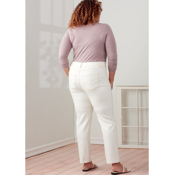 A person with curly hair, wearing a lavender long-sleeved top and white Misses and Women's Vintage Jeans (Buttons/Zipper) by Super Cheap Fabrics' Simplicity - S9266 collection, is standing and facing away from the camera in a bright room with wooden flooring. There is a window frame leaning against the wall in the background.