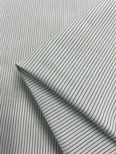 The Shirting - Harp - 145cm from Super Cheap Fabrics features a neatly folded white polyester cotton fabric with thin green pinstripes spaced evenly, creating diagonal lines. This lightweight fabric is known for its smooth surface texture, emphasizing the crisp and clean pattern.