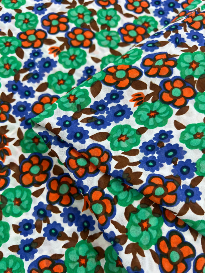 A close-up of the Salt Shrunk Print - Kaleid from Super Cheap Fabrics shows an elegant, vibrant floral pattern with large green and brown flowers, smaller blue flowers, and orange accents on a white background. The fabric's folds highlight its intricate, vivid design. Width: 155cm.