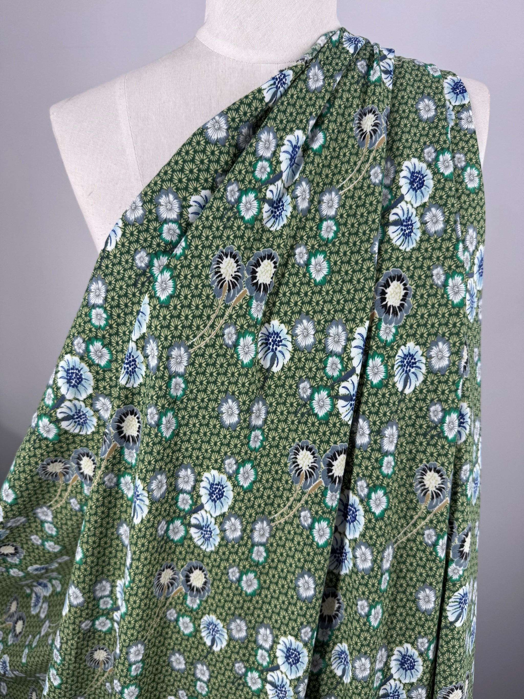 The Printed Crepe - Botan by Super Cheap Fabrics, draped over a mannequin, features an elegant floral pattern with blue, white, and black flowers surrounded by small green leaves on its 148cm wide green fabric.
