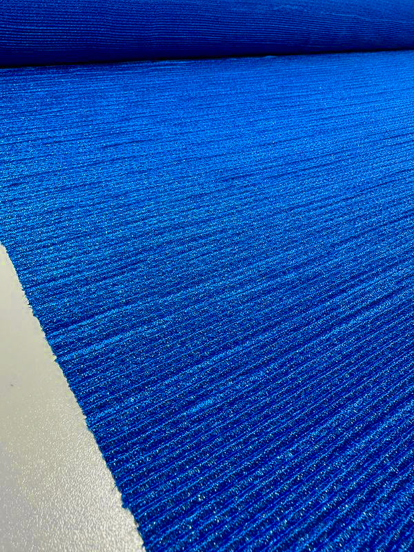 A close-up view of the vibrant Metallic Knit in Cobalt from Super Cheap Fabrics, showcasing its textured, ribbed surface pattern. The medium to heavy weight fabric is stretched out to reveal its full width of 150cm, with the roll of fabric visible in the background. The color is vivid and consistent across the material.