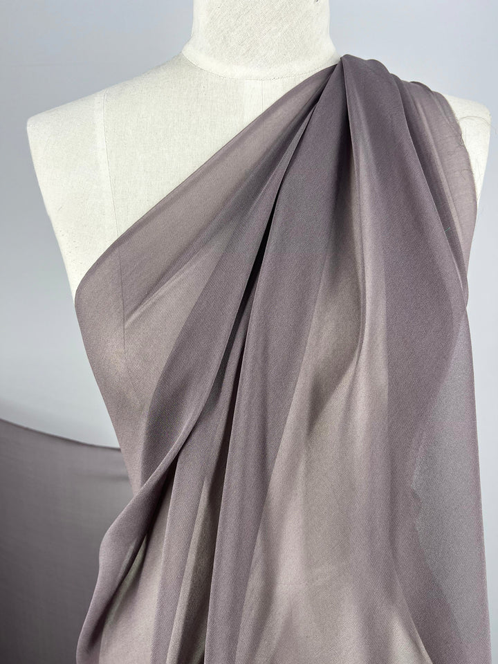 A close-up image shows a draped elegance of Super Cheap Fabrics' "Silk Georgette - Pewter - 135cm" over a dress form mannequin. The lightweight, smooth-textured fabric cascades in gentle, flowing folds, creating soft shadows and highlights. The plain light gray background complements the luxurious 100% silk material perfectly.