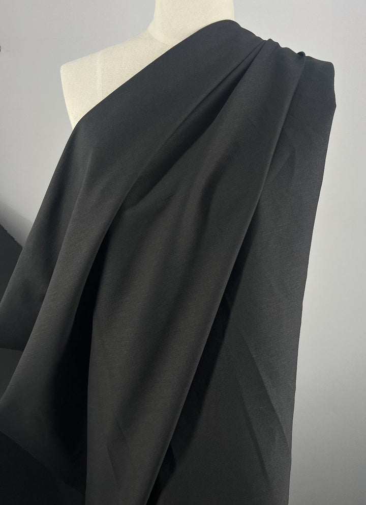 A mannequin is elegantly draped with Super Cheap Fabrics' Blended Viscose Suiting in black, 150cm wide. The lightweight fabric with a subtle sheen cascades gently in drapey folds against a light backdrop.