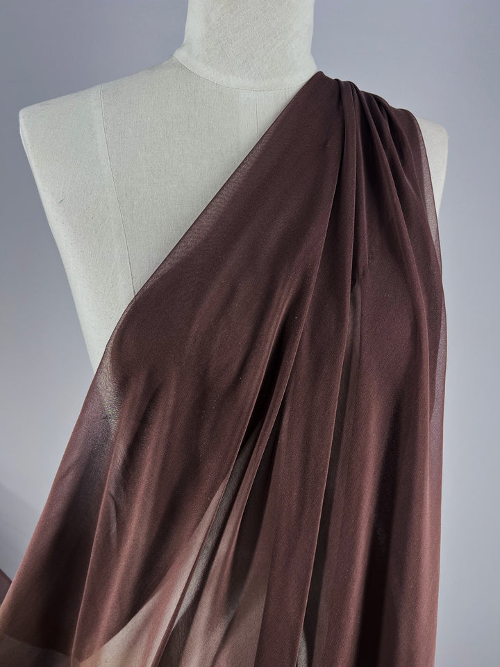 A Super Cheap Fabrics mannequin showcases the Pure Silk Georgette - Chocolate in a sheer, dark brown drape diagonally across the chest, forming elegant folds. This lightweight 135cm fabric is ideal for spring and summer wear, with a gray backdrop enhancing its texture and color.