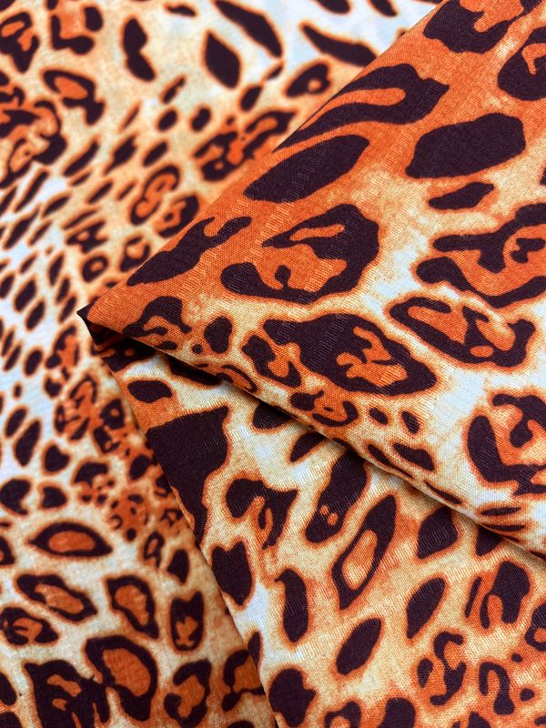 Close-up image of a soft, designer fabric with a vibrant leopard print design from Super Cheap Fabrics. This Textured Deluxe Print - Leo - 155cm features a captivating mix of orange, black, and white colors, creating an authentic wild animal print look. The versatile fabric is folded to showcase the intricate details and texture of the material.