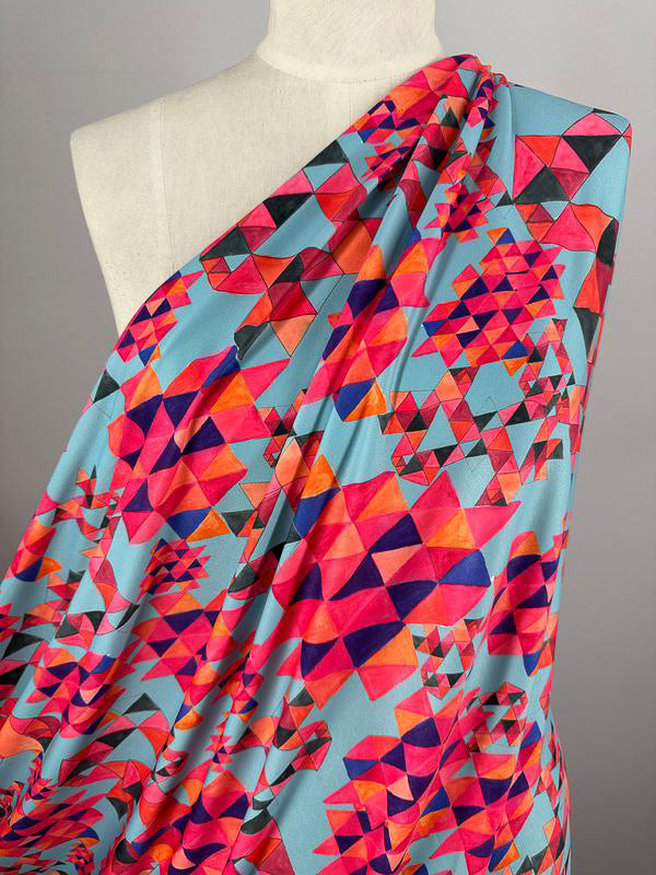 A mannequin showcased in the vibrant "Printed Lycra - Geo Village - 150cm" fabric from Super Cheap Fabrics. This medium-weight, Polyester/Spandex material features an array of red, pink, orange, blue, and black triangles intricately overlapping against a light blue background, creating a dynamic and colorful geometric pattern.