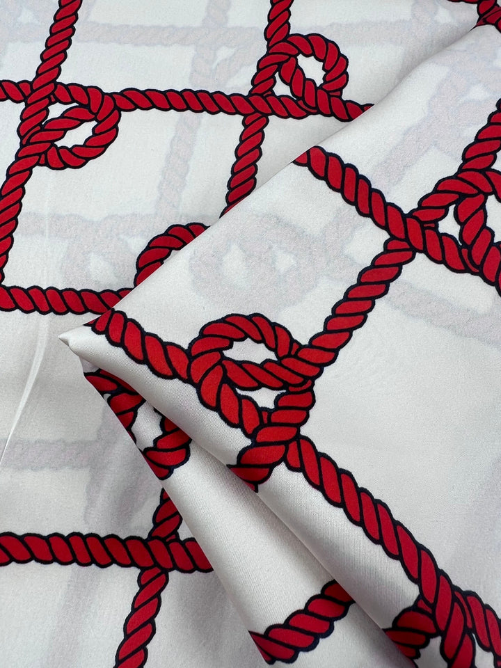 The "Printed Satin - Cruise - 150cm" by Super Cheap Fabrics showcases an elegant red rope pattern on a white background, forming a geometric grid with circular knots. Slight folds add texture, evoking a refined border print design.