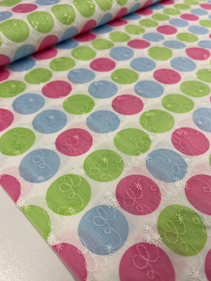 A close-up view showcases the Embroidered Printed Cotton - Candy - 112cm from Super Cheap Fabrics, featuring a vibrant pattern of green, blue, and pink circles enhanced with subtle embroidered swirl designs. Natural cotton fibers spread out on a flat surface to highlight its striking design, making it perfect for both clothing and household items.