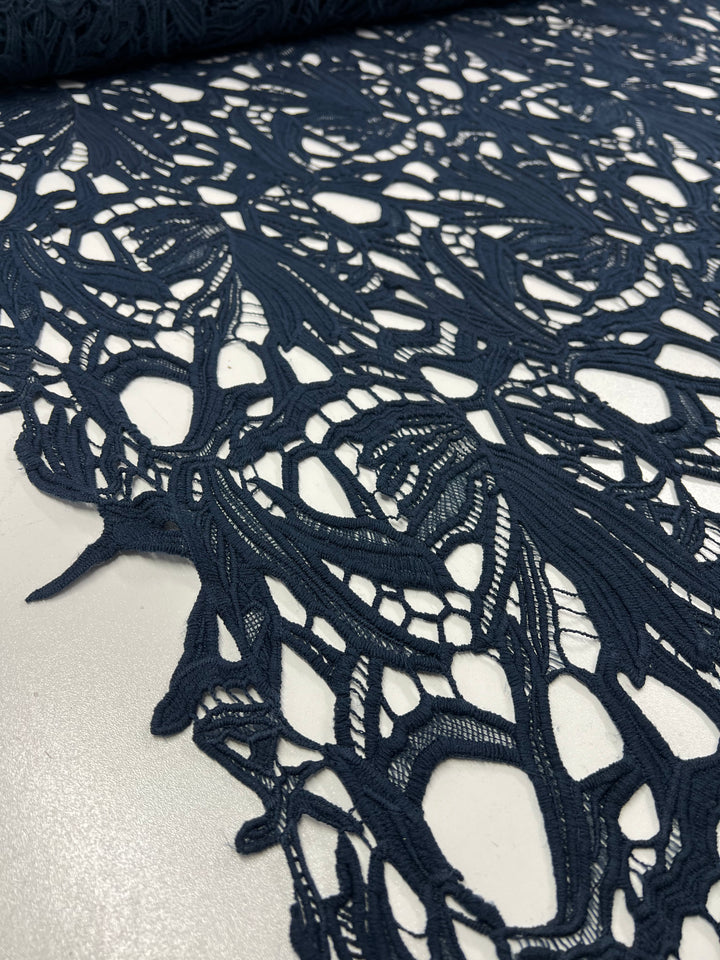 Close-up image of dark blue intricately patterned **Evening Lace - Navy - 100cm** fabric by **Super Cheap Fabrics** spread over a white surface. The lightweight polyester design features various curving and intersecting shapes, creating a delicate and elaborate texture.