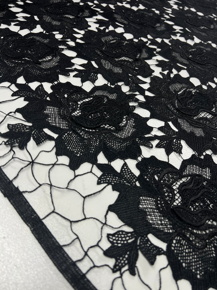 A close-up of intricate black lace fabric featuring a floral pattern of roses and leaves. The 100% polyester design is set against a white background, highlighting the delicate details and elegant texture. This exquisite black fabric, Designer Anglaise Lace - Black - 120cm by Super Cheap Fabrics, boasts a generous 120cm width, perfect for various projects.