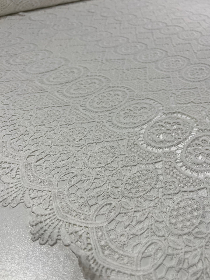 Close-up of an intricate lace fabric with a delicate, detailed pattern featuring floral and geometric designs. The ivory fabric is 116 CM wide and has a soft, elegant texture. The edges have a scalloped, lace trim made from high-quality polyester. This is the Designer Anglaise Lace - Guipure - 116cm by Super Cheap Fabrics.