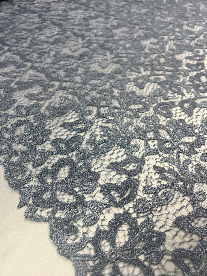 Close-up of a delicate lace fabric with intricate floral patterns. The Lightweight Super Cheap Fabrics Evening Lace - Metallic Blue - 120cm is spread out on a white surface, showcasing its fine details and craftsmanship.