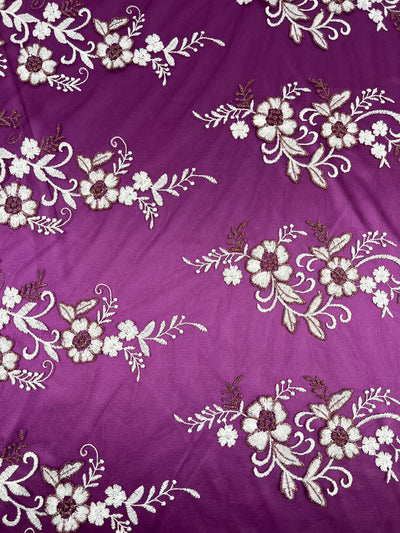 A detailed close-up of the "Embroidered Lace - Plum - 150cm" by Super Cheap Fabrics showcases a beautiful embroidery pattern featuring white and brown flowers with green stems and leaves on a rich plum, lightweight fabric background. The flowers and leaves are intricately arranged in repeated clusters across the polyester fabric.