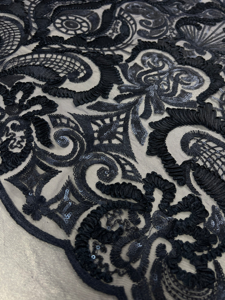 Close-up of an intricately patterned black lace fabric. The design features various motifs including floral and swirl shapes, some areas adorned with small sequins that add a subtle sparkle. This 100% polyester fabric, Evening Lace - Night Shade - 150cm by Super Cheap Fabrics, has a light beige background material, providing contrast to the dark lace.