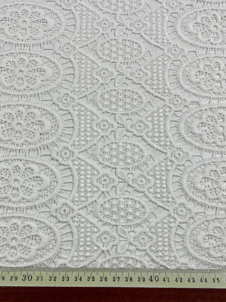 A close-up image of Designer Anglaise Lace - Guipure - 116cm by Super Cheap Fabrics spread out on a flat surface. The polyester lace is intricately patterned with floral and circular motifs. A yellow measuring tape with centimeter markings, reading up to 116 CM, is placed at the bottom edge of the fabric.