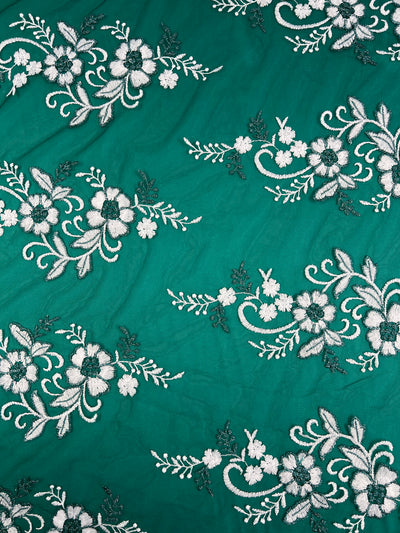 The Embroidered Lace - Aztek by Super Cheap Fabrics features intricate white and dark green floral embroidery on a green fabric, reminiscent of French elegance, with swirling leaves and flowers creating a decorative, elegant design across the 150cm material.