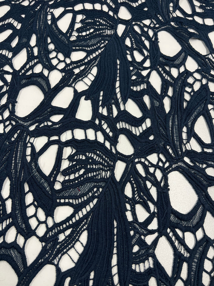 Close-up image of Super Cheap Fabrics' Evening Lace - Navy - 100cm intricate polyester lace fabric with floral and leaf patterns. The lightweight fabric has an open, airy design with solid and sheer material creating a contrast against a white background.