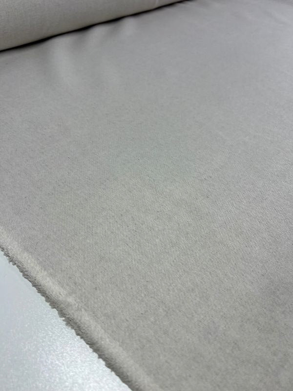 A close-up image displays a roll of Super Cheap Fabrics' Wool Cashmere - Speckley Snow - 150cm, featuring a smooth yet slightly textured surface in light grey or beige. The fabric is spread out flat on a white surface, with the edge of the roll visible in the background.