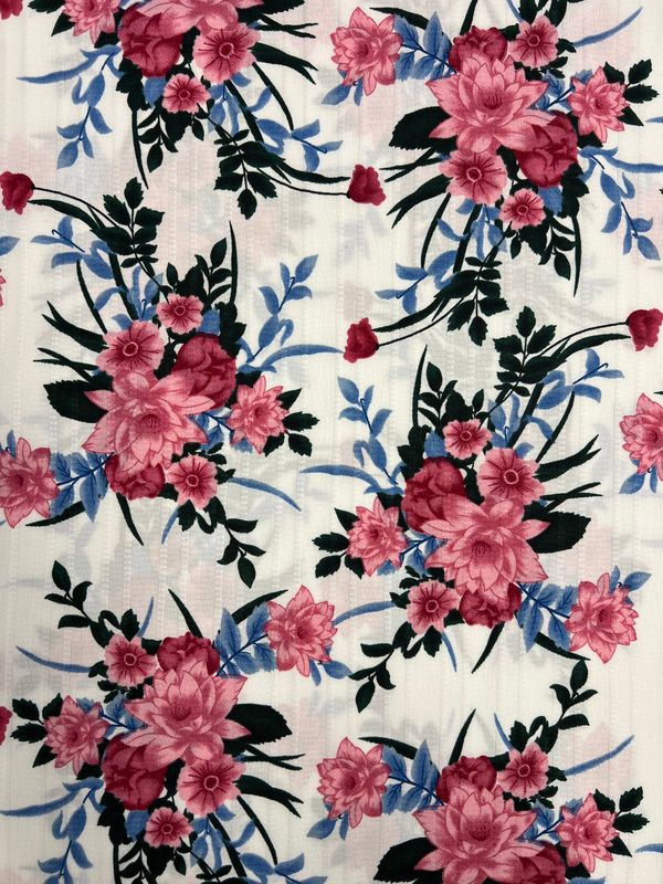 The "Textured Deluxe Print - Catch The Bouquet - 155cm" by Super Cheap Fabrics features a floral pattern with pink and red flowers, green leaves, and blue accents on a white background. Its symmetrical design offers vibrant, eye-catching repetition, making it versatile for various uses.