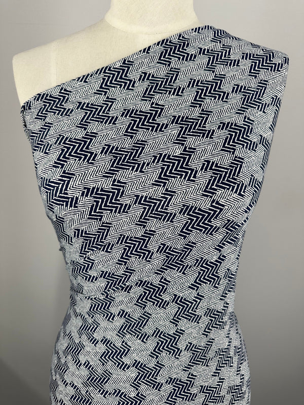 A close-up image of a one-shoulder mannequin draped in Super Cheap Fabrics' Printed Lycra - Tazo - 150cm. The intricate blue-and-white zigzag pattern, crafted from medium-weight Polyester/Spandex fabric, features alternating directional zigzags, creating a visually dynamic and textured appearance. The plain background emphasizes the fabric's detail.