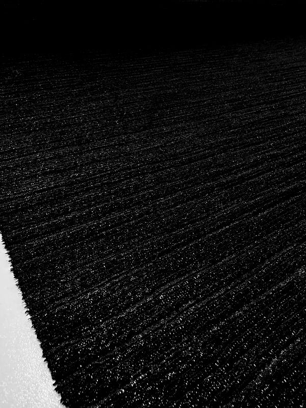 A close-up photograph of the Super Cheap Fabrics' Metallic Knit in Black (150cm), showcasing its textured, medium to heavy weight knit similar to carpet. The image accentuates the fibrous material and the subtle sheen on its surface, with a hint of light-colored flooring visible at the edge. The lighting creates a striking contrast between light and shadow.