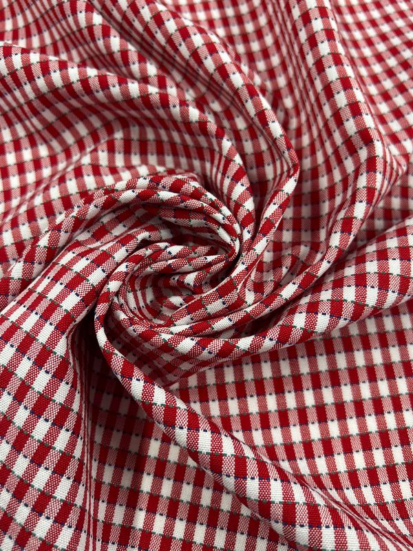 Close-up of the Shirting - Riding by Super Cheap Fabrics, 145cm wide red and white checkered polyester fabric swirled into a spiral. The lightweight material showcases a classic gingham pattern with smooth texture and gentle folds forming around the center swirl.