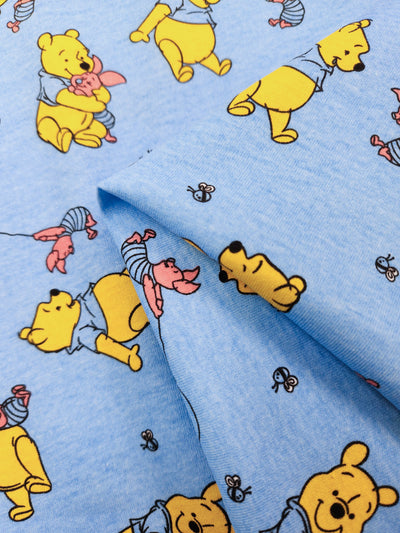 The Super Cheap Fabrics Printed Jersey - Winnie & Co - 170cm features charming illustrations of a playful yellow bear, often with a balloon, and a pink pig in various poses. Both wear shirts on a light blue background. Made from poly/cotton jersey, it combines comfort and style.