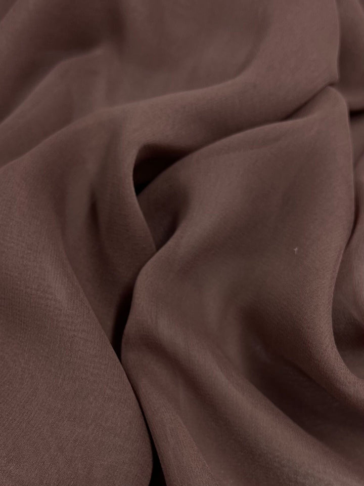 A close-up image of Super Cheap Fabrics' Silk Georgette - Shroon (135cm) reveals the textured brown fabric with soft folds and creases, creating a flowing, wavy pattern across the lightweight material. The surface appears smooth and slightly reflective, capturing light and shadow to highlight its intricate details—perfect for spring and summer outfits.