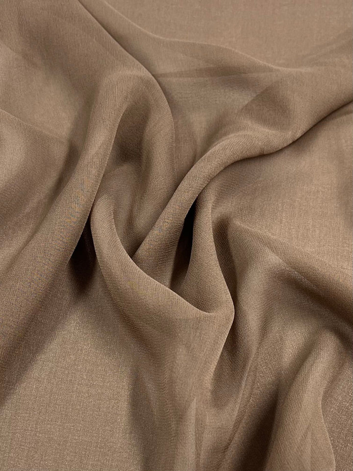 A close-up of the "Silk Georgette - Bark" fabric from Super Cheap Fabrics shows its beige, lightweight material with slight wrinkles and folds. The fabric appears soft and smooth, draping gently to give it a sense of fluidity and texture, making it an ideal choice for spring and summer outfits or home decor items. This silk georgette is available in a 135cm width.