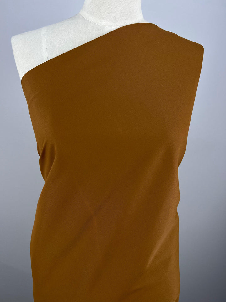 A mannequin is draped in a single-shoulder Delustered Satin - Caramel Cafe - 150cm by Super Cheap Fabrics, covering one shoulder completely and leaving the other uncovered. The background is plain gray, focusing attention on the luxurious fabric.