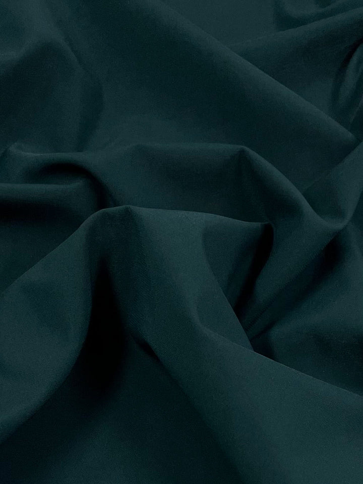 A close-up of dark green, lightweight Delustered Satin - Ponderosa Pine - 150cm by Super Cheap Fabrics with soft folds and wrinkles. The surface texture appears smooth and slightly matte, with shadows highlighting the creases and draping, indicating its 100% polyester composition.