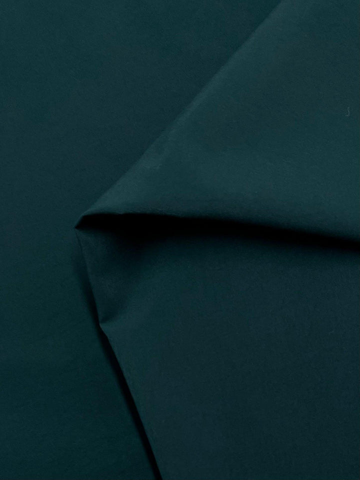 A close-up image of a dark green satin fabric with a smooth texture. The Delustered Satin - Ponderosa Pine - 150cm by Super Cheap Fabrics is folded at the top left, creating a visible crease and shadow, adding depth to the image. The overall appearance of this lightweight fabric is clean and uniform.