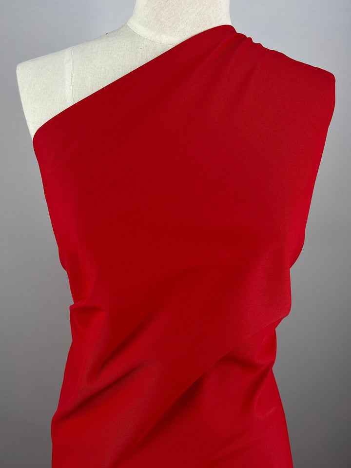 A Super Cheap Fabrics Delustered Satin - Goji Berry - 150cm, one-shoulder dress draped elegantly on a white mannequin against a plain gray background. The 100% polyester satin fabric appears smooth and slightly glossy, creating a flowing design across the mannequin's form.