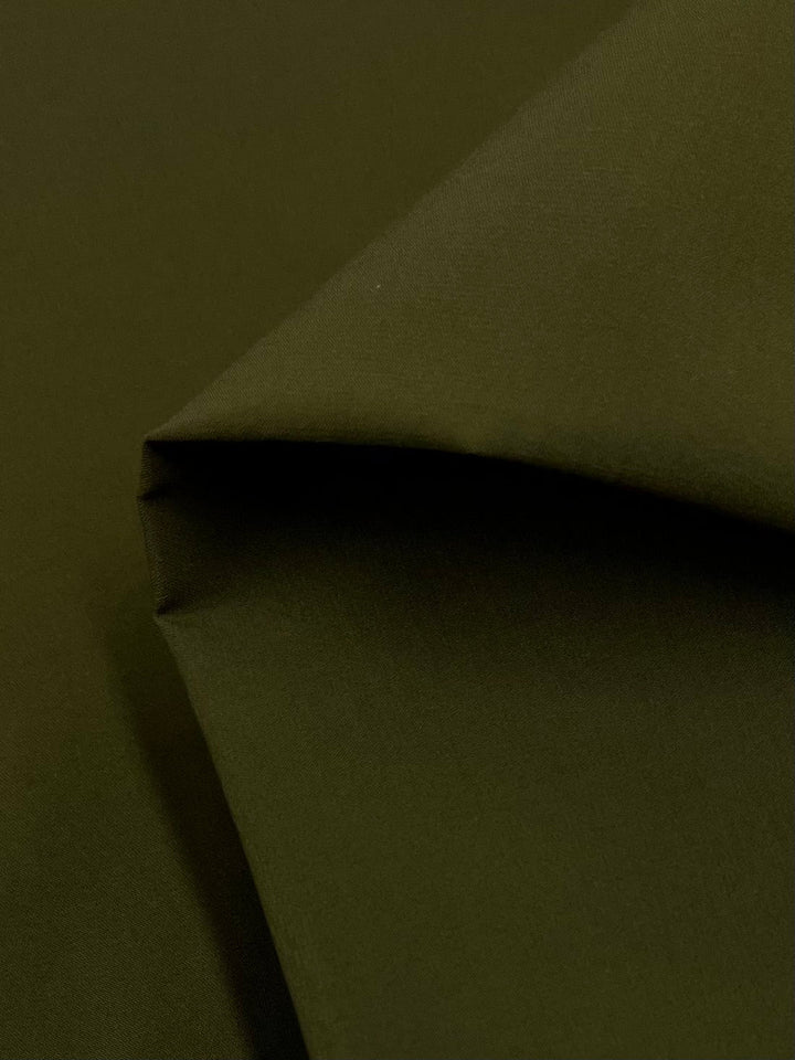 A close-up view of a folded piece of Delustered Satin - Olive Drab - 150cm with a smooth texture from Super Cheap Fabrics. The 100% polyester material is layered over itself, showing the corner fold prominently. The lighting highlights the shape and color of the satin-finish fabric.