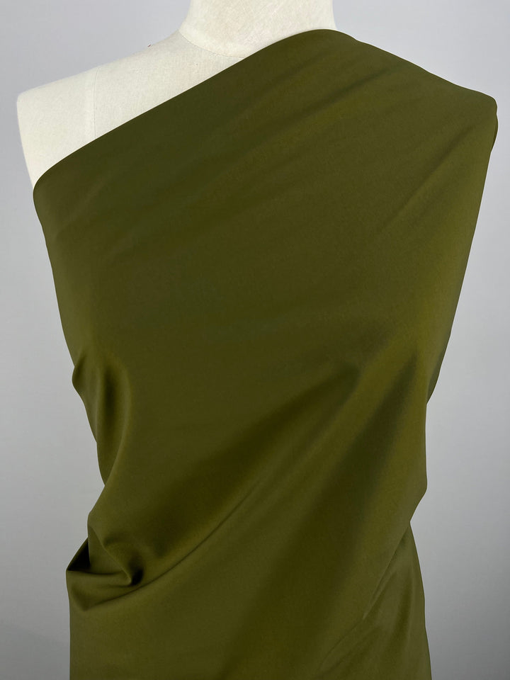 A mannequin draped with Delustered Satin - Olive Drab - 150cm from Super Cheap Fabrics, styled in a one-shoulder design. The smooth, soft 100% polyester material has a slight sheen and drapes elegantly over the mannequin, showcasing its texture and flow. The background is plain and light-colored, keeping the focus on the fabric.