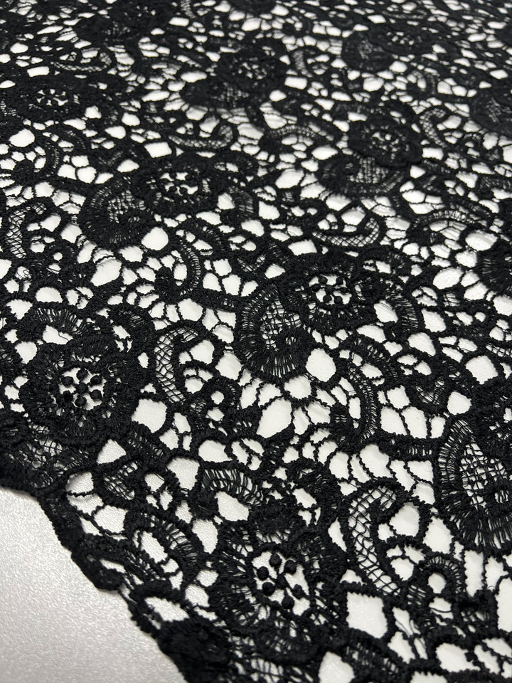 A close-up of a piece of elegant Anglaise Lace - Black Garden - 115cm by Super Cheap Fabrics with intricate floral and geometric patterns. The versatile lace showcases various shapes and textures, set against a light background that highlights its ornate features.