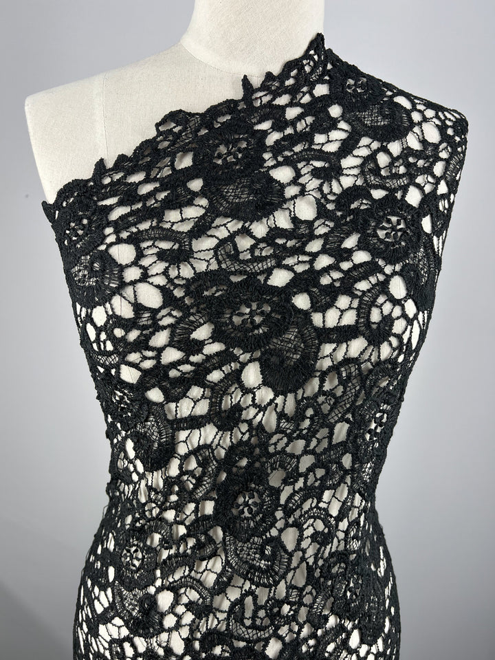 A mannequin displays a versatile one-shoulder garment made of intricate Anglaise Lace - Black Garden - 115cm from Super Cheap Fabrics with floral patterns. The design features semi-sheer areas with detailed, intertwining motifs, creating an elegant and sophisticated look against a light background.