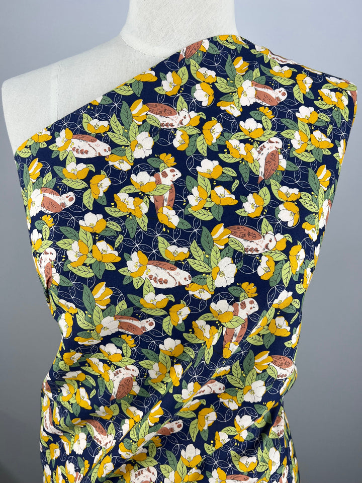 A mannequin is draped in a dark blue cotton fabric adorned with a vibrant botanical print of sloths hanging among yellow flowers and green leaves. Wrapped diagonally across the mannequin, this versatile fabric showcases the colorful and whimsical design, perfect for multiple uses. The Printed Cotton - Wild Camellias - 150cm from Super Cheap Fabrics brings an enchanting touch to any project.