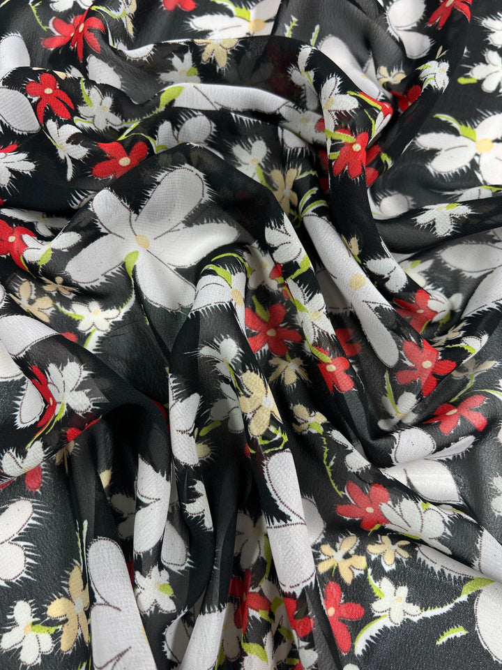 A close-up of the "Printed Georgette - Jasmine Cluster - 150cm" fabric by Super Cheap Fabrics. The sheer polyester material features a floral pattern with a black background adorned with white, red, and yellow painted flowers, creating a vibrant and lively design. The folds and texture of the georgette fabric are also visible, giving it a dynamic appearance.