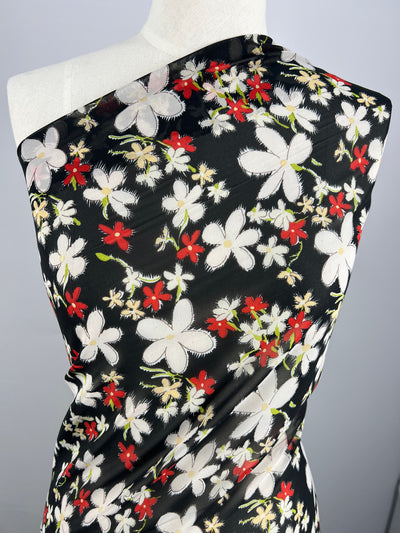 A mannequin is draped in Super Cheap Fabrics' Printed Georgette - Jasmine Cluster - 150cm, adorned with painted flowers. The white, red, and pale yellow hues create a vibrant contrast against the dark background. The sheer polyester fabric is placed asymmetrically, highlighting its delicate design.