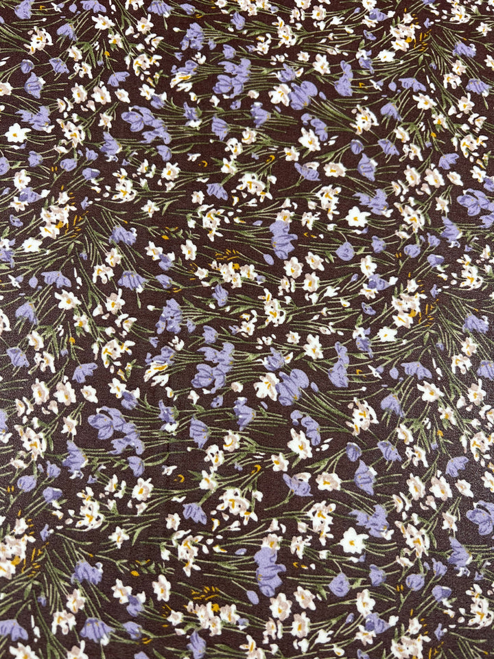 Printed Georgette - Floral Strokes - 150cm from Super Cheap Fabrics showcases a delicate floral pattern with elegant purple and white blooms alongside green stems and leaves, all spread across a dark background. The petite flowers are scattered intricately, creating a dense, intricate design on sheer Georgette fabric.