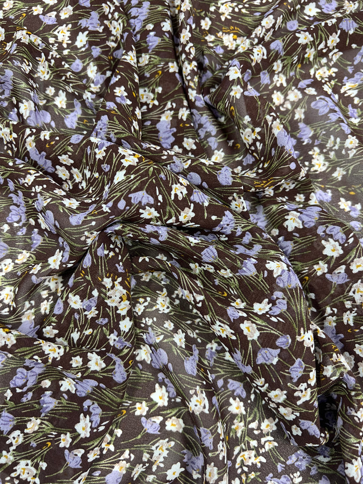 Here is a close-up of Super Cheap Fabrics' Printed Georgette - Floral Strokes - 150cm, showcasing an elegant pattern of small white and light purple flowers with green stems on a dark background. The sheer fabric is slightly crumpled, beautifully displaying its texture and intricate print details.