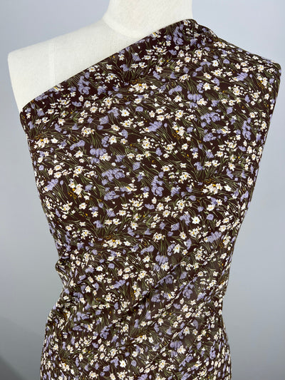 A close-up of a mannequin draped in Super Cheap Fabrics' Printed Georgette - Floral Strokes fabric, featuring a delicate dark brown material adorned with small light purple flowers and subtle white accents. The sheer Georgette is styled in an asymmetrical, one-shoulder design, complete with soft folds and a fitted appearance.