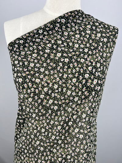 A mannequin draped in Super Cheap Fabrics' Printed Georgette - Dark Blooming Seeds, a black one-shoulder fabric adorned with small white and pink flowers. The sheer polyester hangs smoothly, displaying its painted floral design against a light gray background.