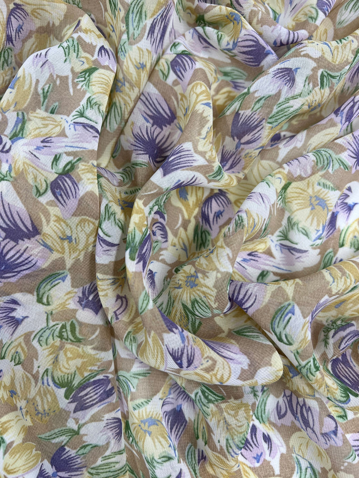 A close-up of the Super Cheap Fabrics' Printed Georgette - Earths Bloom - 150cm showcases its delicate, crumpled texture and floral pattern. This design features painted flowers in shades of yellow, purple, and green set against a light brown background. The soft texture of the sheer polyester fabric enhances the vibrant colors, creating a lively and dynamic appearance.