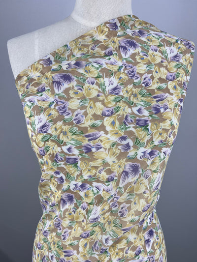 A dress on a mannequin featuring a single-shoulder design and covered in painted flowers in purple, yellow, white, and green leaves against a beige background. The "Printed Georgette - Earth's Bloom - 150cm" from Super Cheap Fabrics offers a smooth, silky texture that fits snugly on the mannequin.